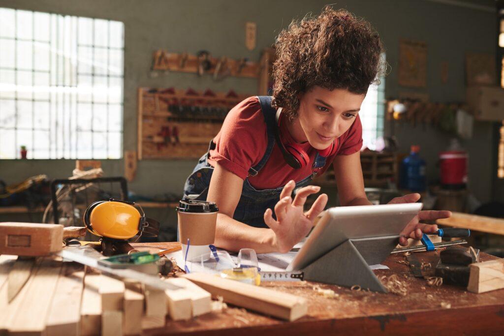 Junge Frau in einer Werkstatt, die an einem Tablet arbeitet. Umgeben von Holz, Werkzeugen und Arbeitsmaterialien plant sie ein Projekt für nachhaltige Massivholzmöbel.