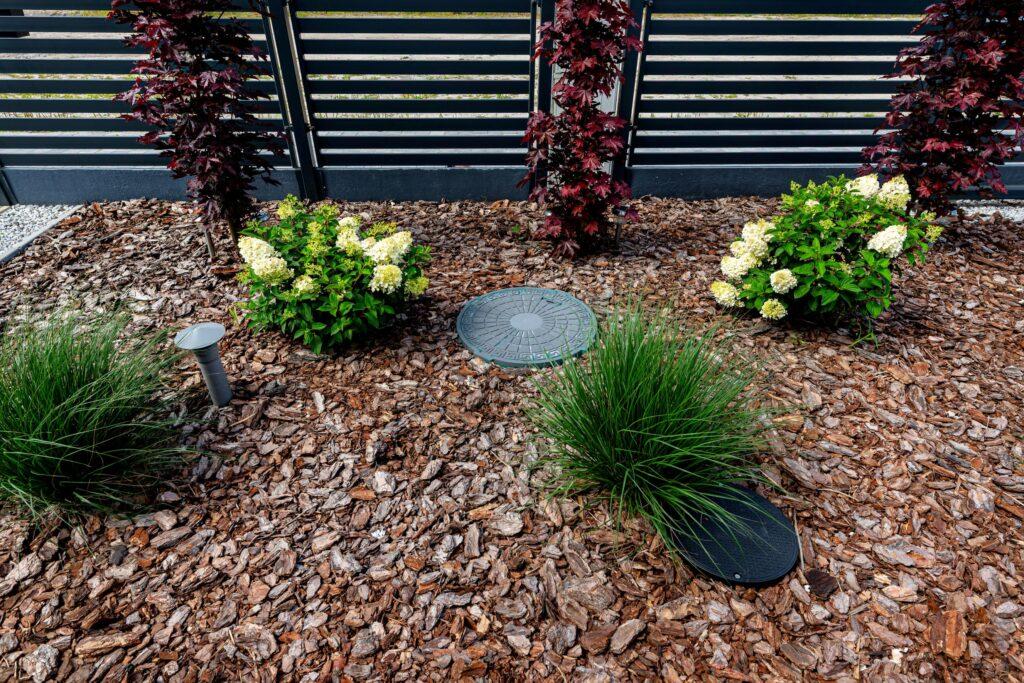 Abwassertank im Garten stilvoll integriert unter Mulch und zwischen Ziersträuchern vor moderner Sichtschutzwand